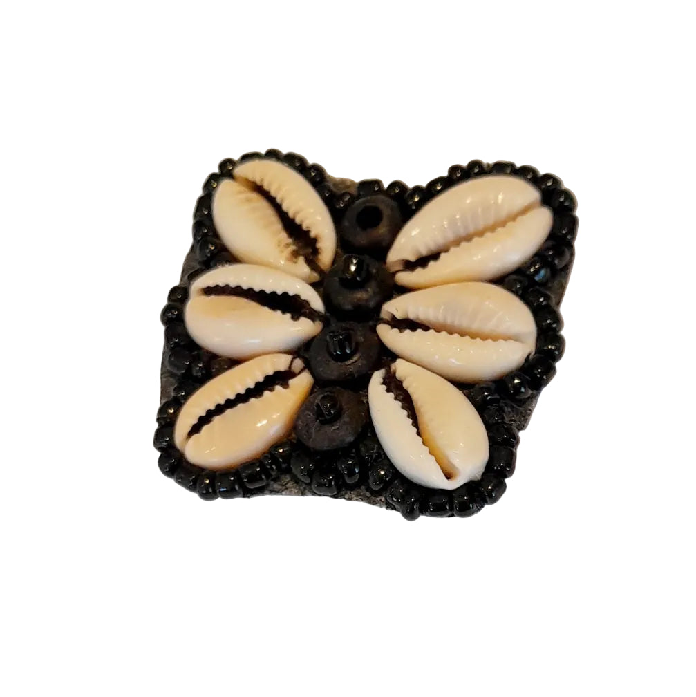 A square black decorative pendant featuring six cowrie shells arranged in a flower pattern, with small black beads around the borde